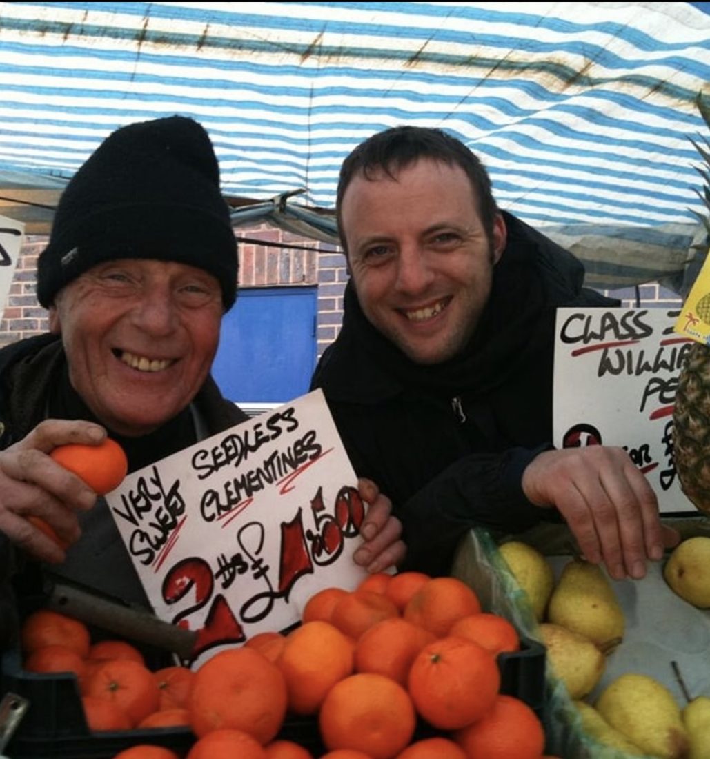 Two fruit traders grinning behind a pile of clementines